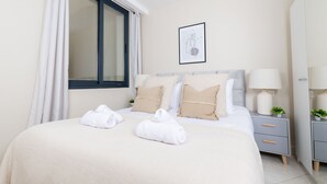 Una cama bien hecha con dos toallas, una mesita de noche con lámpara y una pequeña planta.