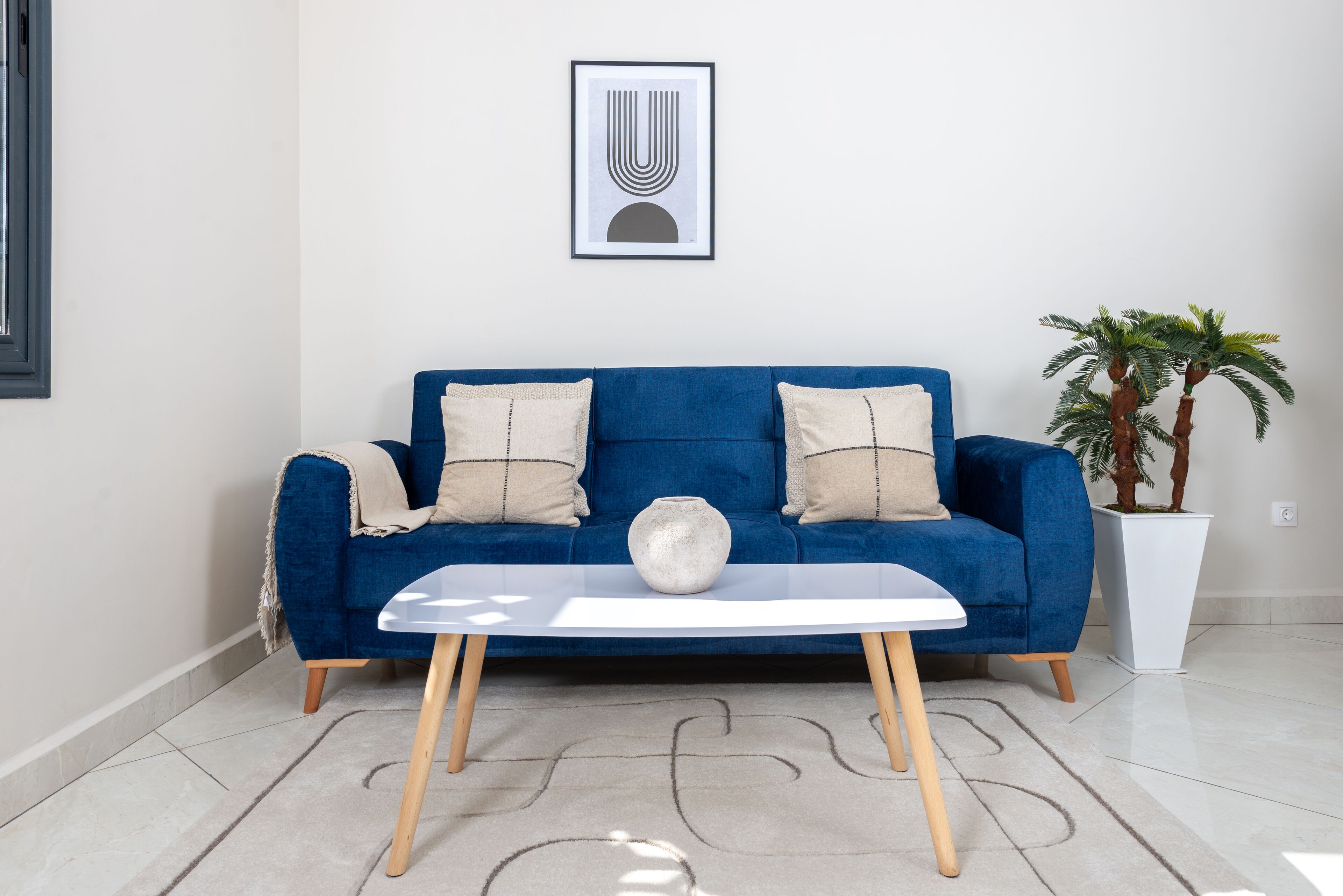 Een blauwe bank met twee beige kussens, een witte vaas en een lijst met abstracte kunst aan de muur.