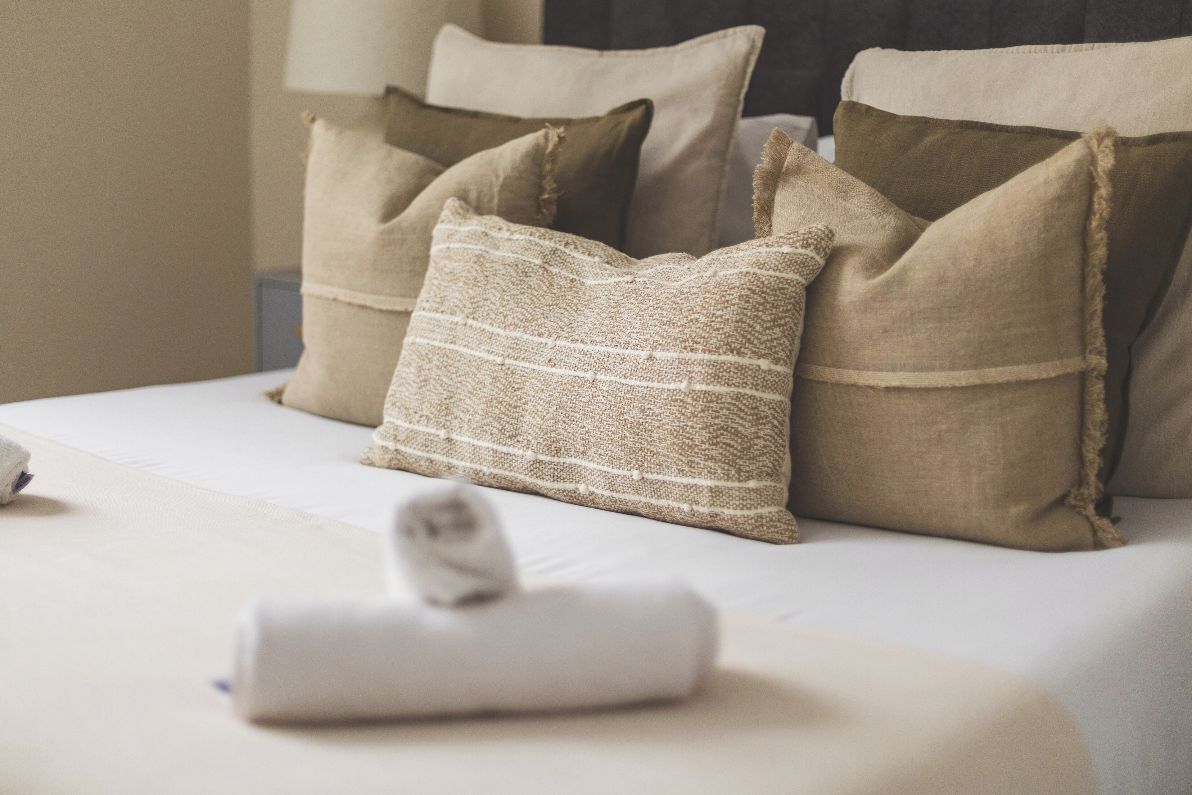 A neatly made bed with beige and brown pillows, a rolled white towel, and a lamp in the background.