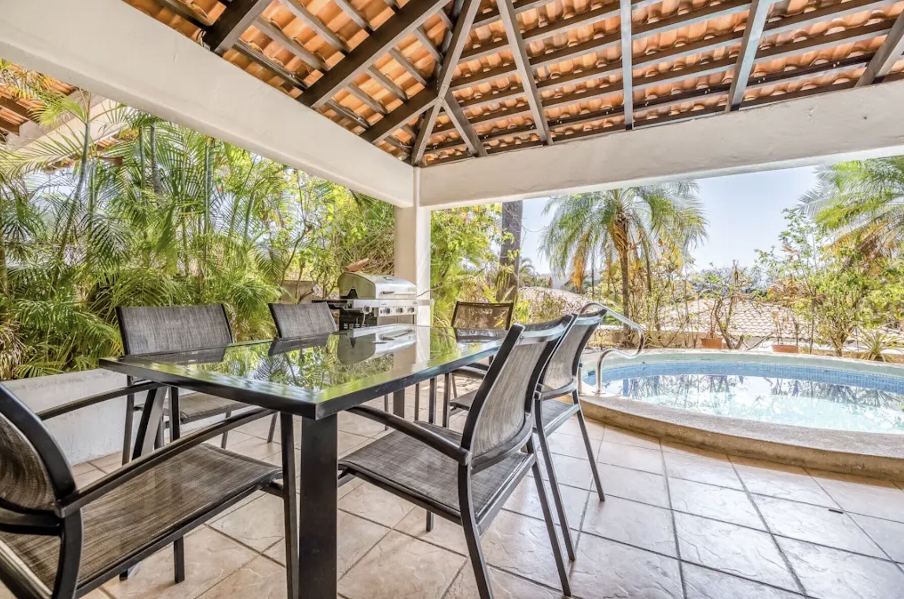 A covered outdoor area with a glass table and chairs, a grill, and a pool.