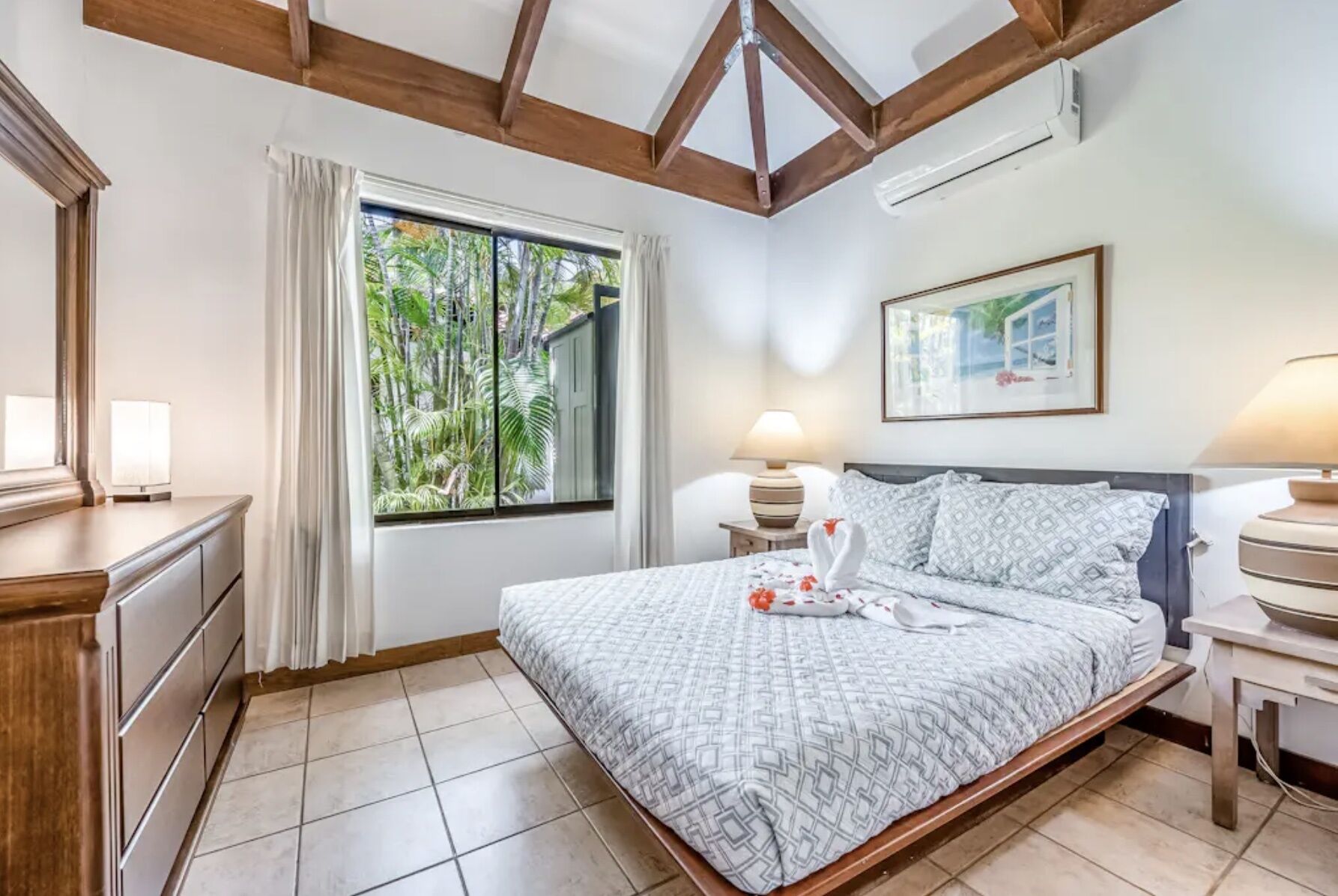 A hotel room with a large bed, a window with curtains, a bedside table with a lamp, and a framed picture on the wall.