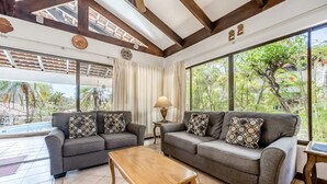 A living room with two grey sofas, a wooden coffee table, and a large window offering a view of greenery. - Beautiful 3 bedroom and 2 bathrooms villa close to beach. (Playa Hermosa)