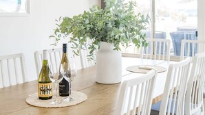 A dining table with two wine bottles, a vase with green foliage, and white chairs. - Strandhytta 10 Cant Rd Goolwa South (Goolwa South)