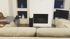 Un salon avec un canapé beige, une cheminée et un téléviseur posé sur un meuble. Des étagères ornées d’objets décoratifs sont fixées au mur.