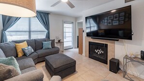Uma sala de estar moderna com sofá seccional, pufe, TV de tela plana, lareira, ventilador de teto e uma grande janela com cortinas.