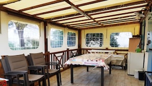 Ein überdachter Patio-Bereich mit Holzboden, einem Tisch mit Blumenmuster-Tischdecke und mehreren Stühlen.