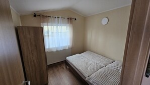 Una habitación pequeña con cama individual, un armario de madera, un radiador y una ventana con cortinas translúcidas.