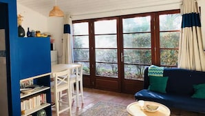 Une pièce avec une bibliothèque bleue, une table blanche avec des chaises, un canapé bleu avec des coussins verts et de grandes fenêtres avec des rideaux.