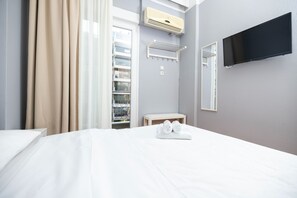 A hotel room with a bed, a TV mounted on the wall, an air conditioning unit, a towel rack, and a window with curtains.