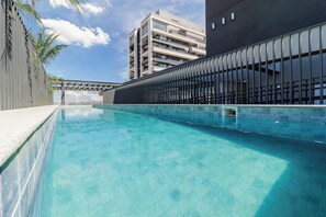 En swimmingpool med kristallklart blått vatten, omgiven av en modern byggnad med balkonger och ett staket av metall.