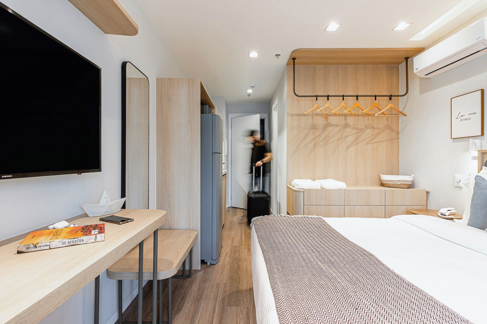 A modern hotel room with a bed, a desk with a chair, a television, and a bathroom with a shower and towel rack.