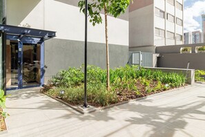 A modern building entrance with a blue canopy, a small garden area with various plants, and a tiled pathway.