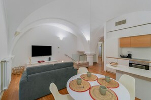 Ein modernes Wohnzimmer mit einem grauen Sofa, einem Fernseher und einem Essbereich mit einem weißen Tisch und vier grauen Tassen.