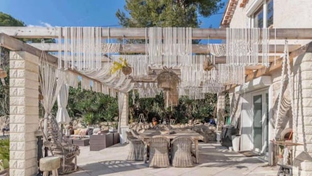 Un patio al aire libre con columnas de piedra, muebles de mimbre y decoraciones colgantes de macramé.