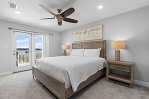 A bedroom with a large bed, ceiling fan, two bedside tables with lamps, a view through a sliding door, and a painting on the wall.