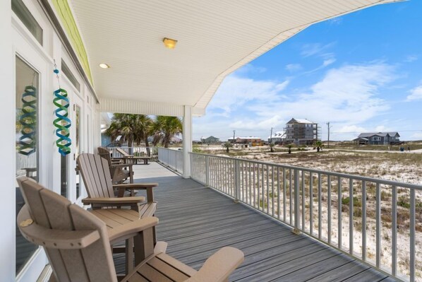 A covered outdoor area with wooden chairs and tables, a white railing, and a view of the beach and buildings in the distance.