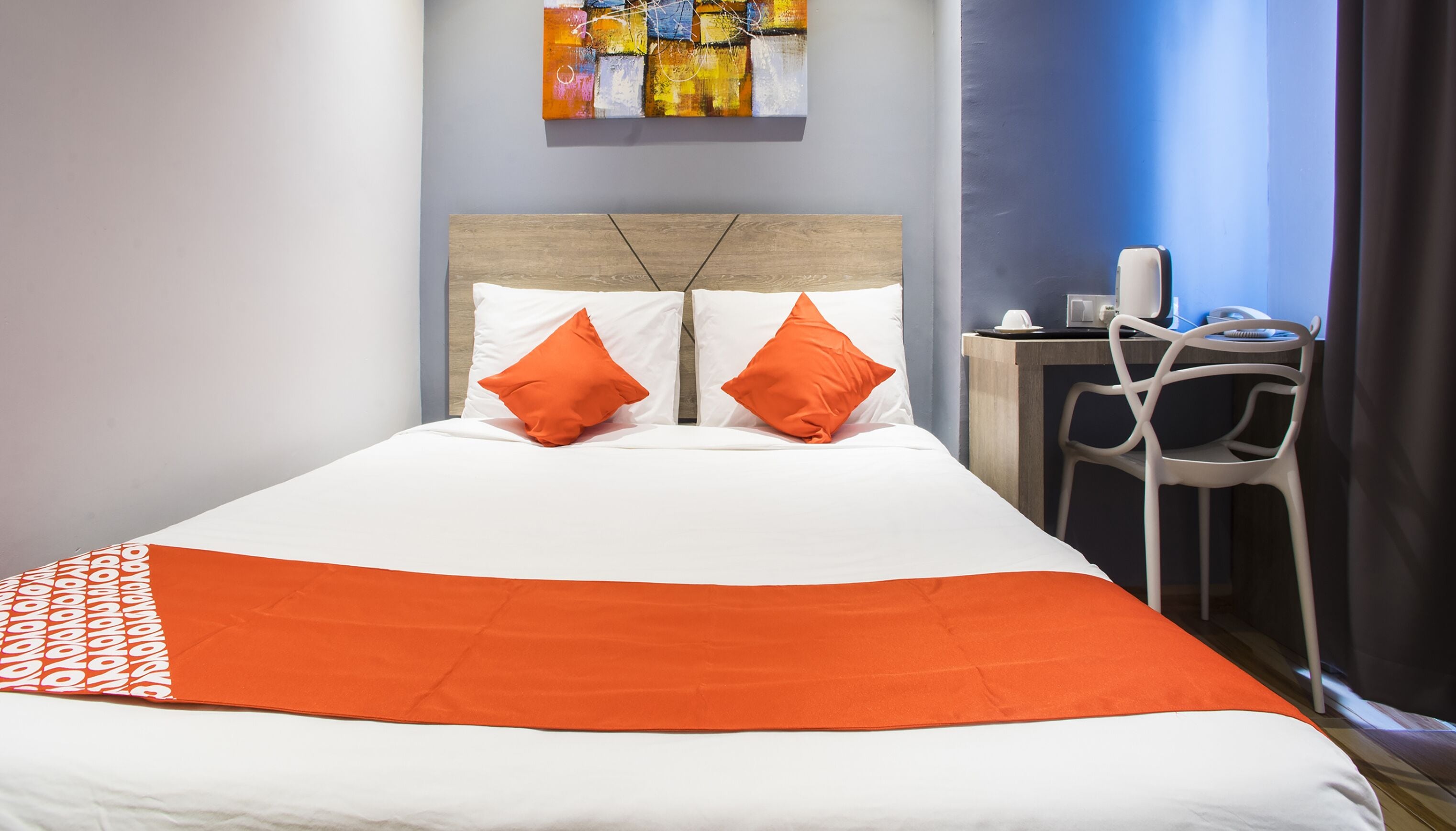 Habitación de hotel con una cama, dos almohadas naranjas, una colcha blanca, un escritorio pequeño con una silla y un cuadro en la pared.