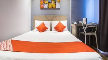 Une chambre d’hôtel avec un lit, deux oreillers orange, un couvre-lit blanc, un petit bureau avec une chaise et un tableau accroché au mur.