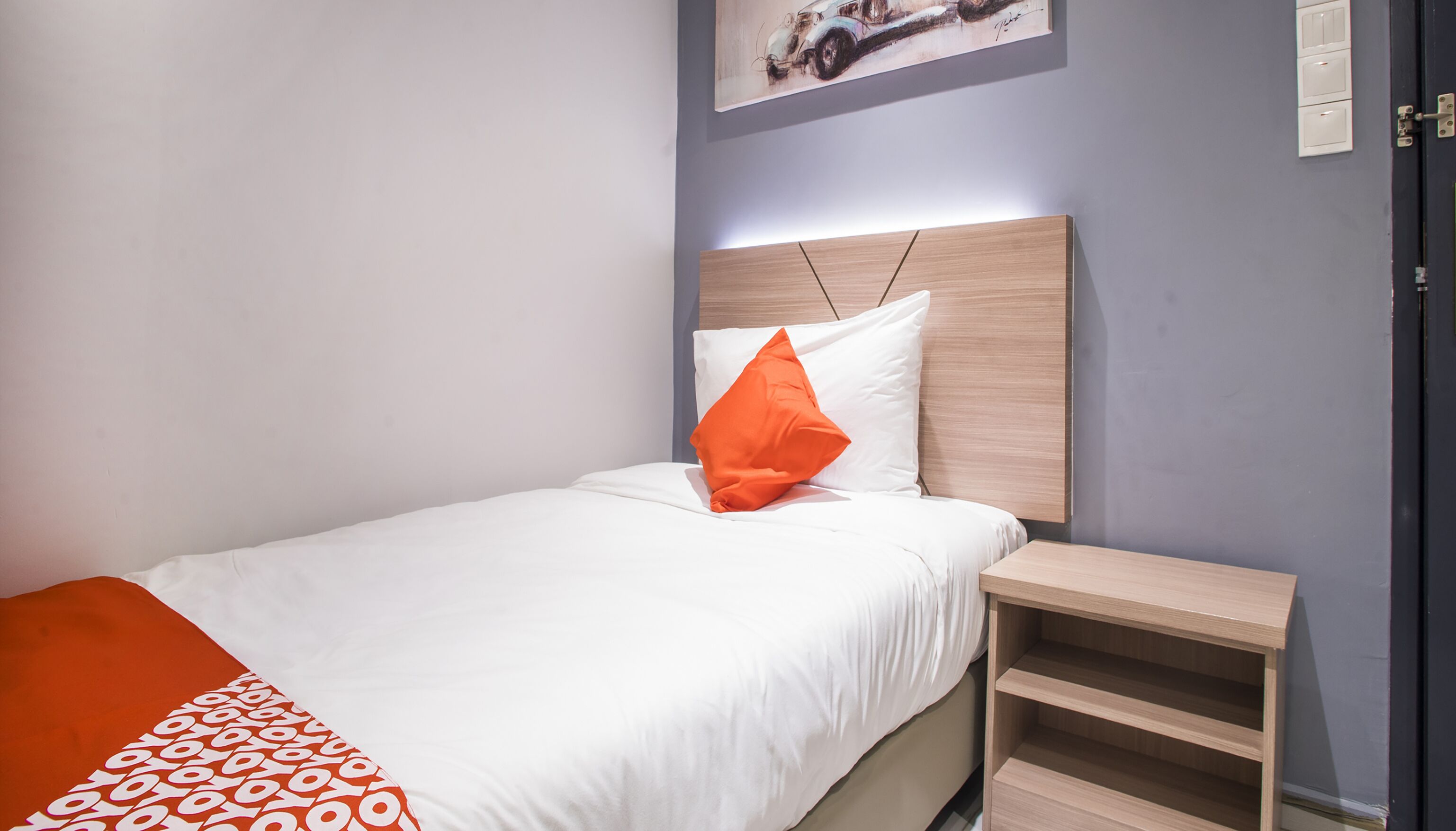A neatly made bed with white bedding and an orange pillow, a wooden nightstand, and a small shelf unit.