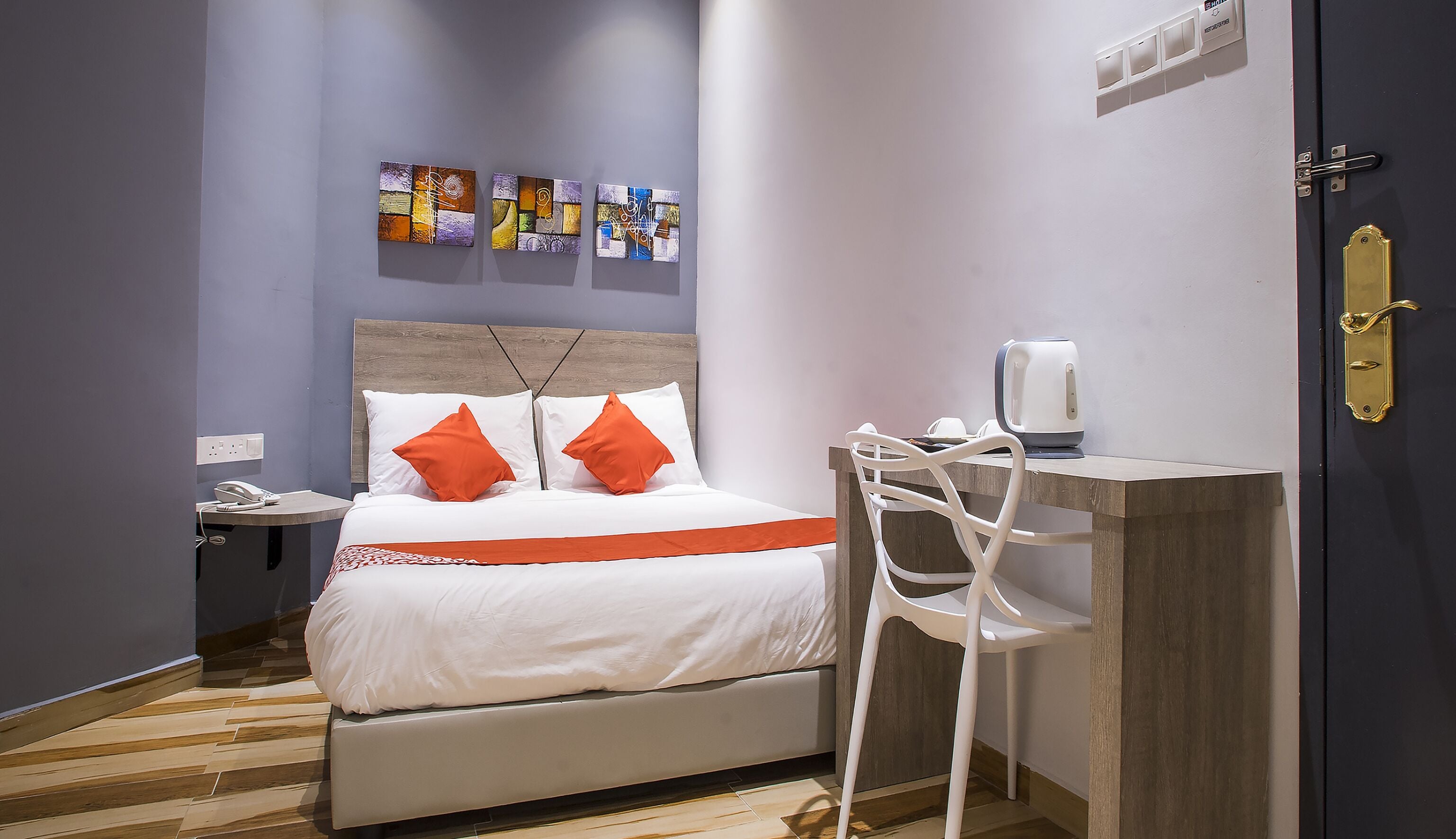 A hotel room with a bed, a bedside table with a phone, a chair, a desk with a coffee maker, and artwork on the wall.