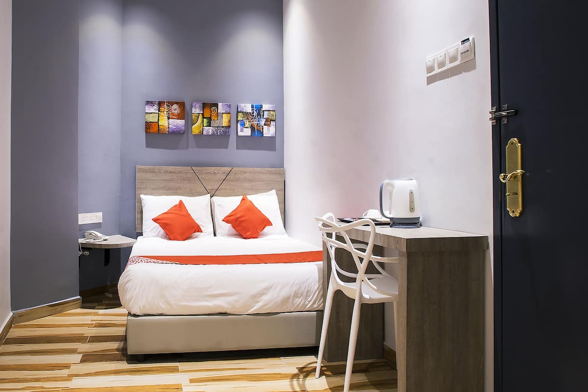 Une chambre d’hôtel étroite, avec un lit, un bureau et une chaise. Des œuvres d’art sont accrochées au mur au-dessus du lit, et il y a une porte avec une poignée.