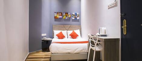 Une chambre d’hôtel étroite, avec un lit, un bureau et une chaise. Des œuvres d’art sont accrochées au mur au-dessus du lit, et il y a une porte avec une poignée.