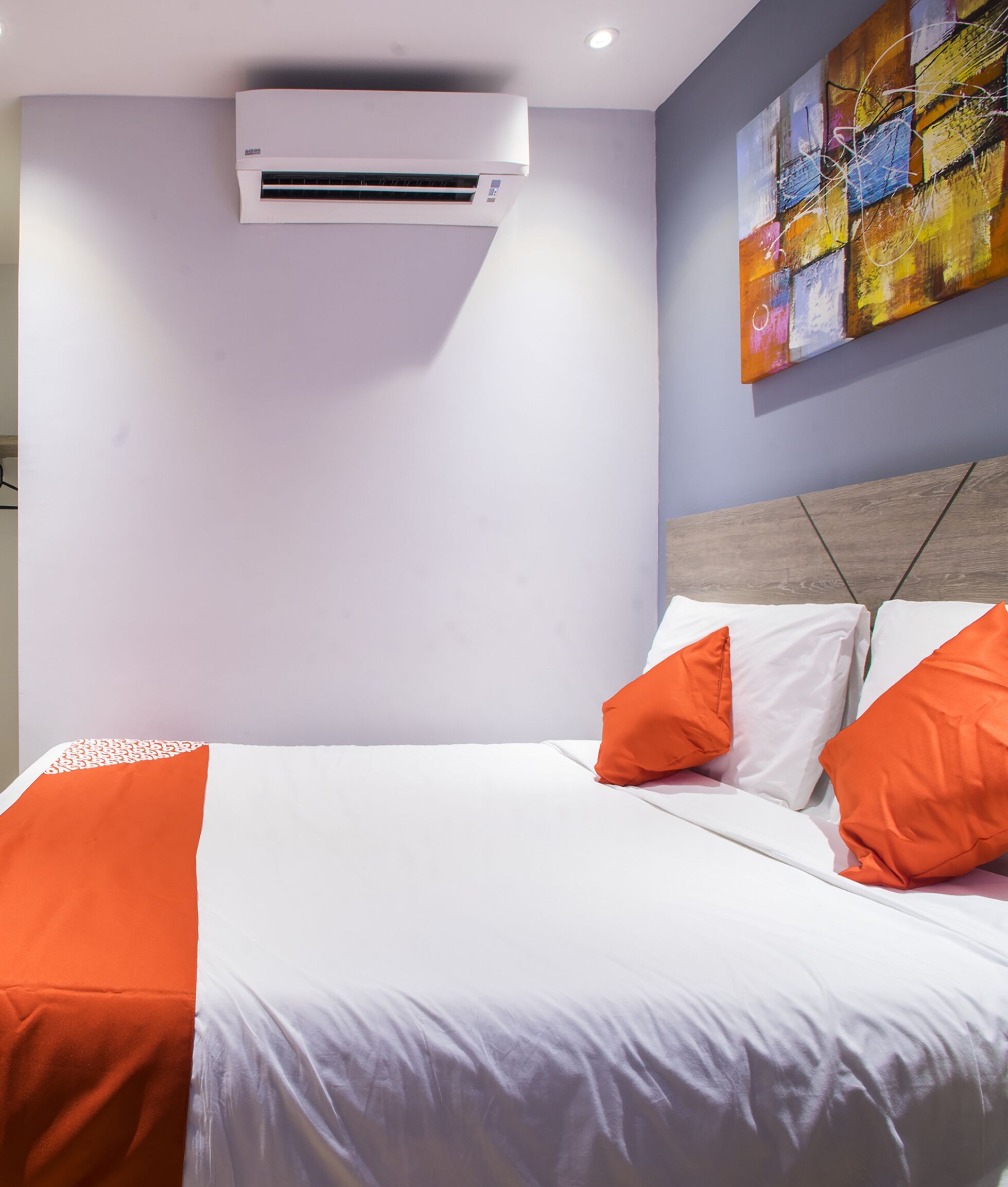 A hotel room with a bed, white bedding, orange pillows, an air conditioning unit, and a colorful abstract painting on the wall.