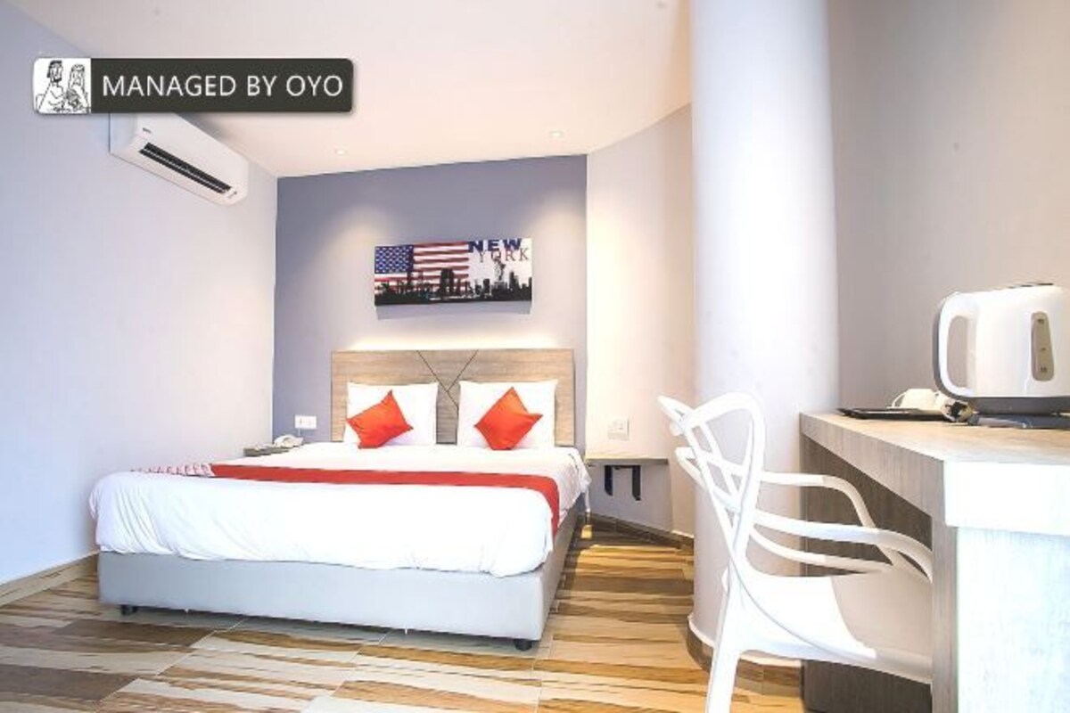 Une chambre d’hôtel comprenant un lit, un bureau, une chaise, un climatiseur et un mur décoré d’une vue sur la ville et d’un drapeau.