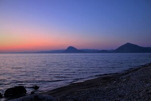 A beach with calm water, pebbly shore, and distant mountains.