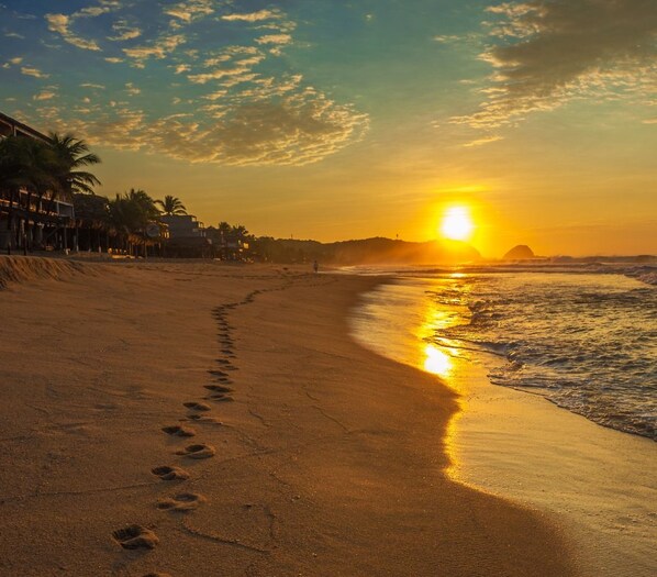 A beach with footprints leading to the water, palm trees, and a sunset in the background. - HOSTAL LEDEZTRUS (San Pedro Pochutla)