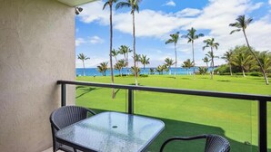 A balcony with a glass table and chairs, overlooking a well-manicured lawn with palm trees and a view of the ocean. - Kihei Surfside #213: Oceanfront Charm and Comfort with Breathtaking Views (Kihei, Maui)
