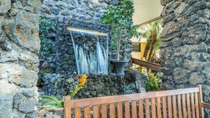 A stone wall with a waterfall feature, a wooden bench, and potted plants. - Kihei Surfside #213: Oceanfront Charm and Comfort with Breathtaking Views (Kihei, Maui)