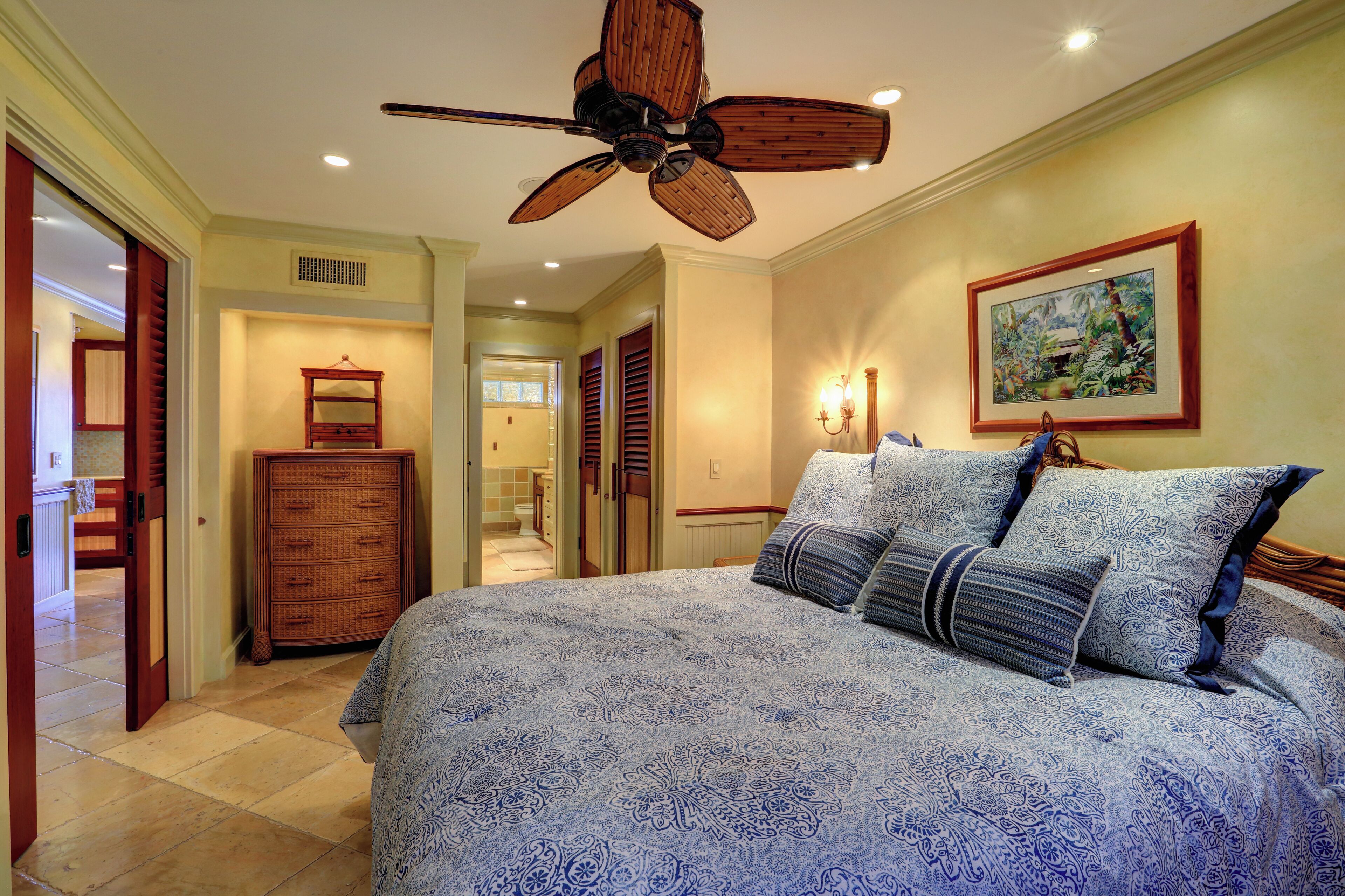 A bedroom with a large bed, a ceiling fan, a dresser, and a painting on the wall.