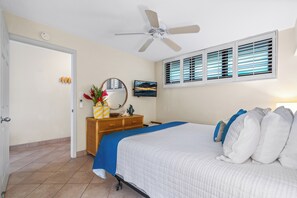 A hotel room with a bed, a ceiling fan, a window with blinds, a wooden nightstand, a TV, and a door. - Kihei Surfside #304 - Spectacular Oceanfront, A/C, 2 Sofa Beds; 1/1.5 baths (Kihei, Maui)