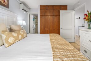 Un dormitorio con cama, armario de madera, cómoda blanca y baño visible a través de una puerta abierta.