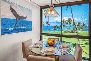 Uma sala de jantar com mesa de vidro, cadeiras e vista para paisagem tropical com palmeiras e campo de golfe.