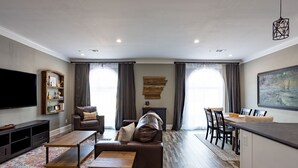 A modern living room with a sofa, coffee table, and TV. There is a dining area with a table and chairs. The room has a large window with curtains and a painting on the wall. - Historic charm Modern Luxury (Batesville)