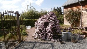 A gravel path leading to a gated entrance, a bush with pink flowers, and a building with a sloped roof. - L'Ermite (Saint-Algis)