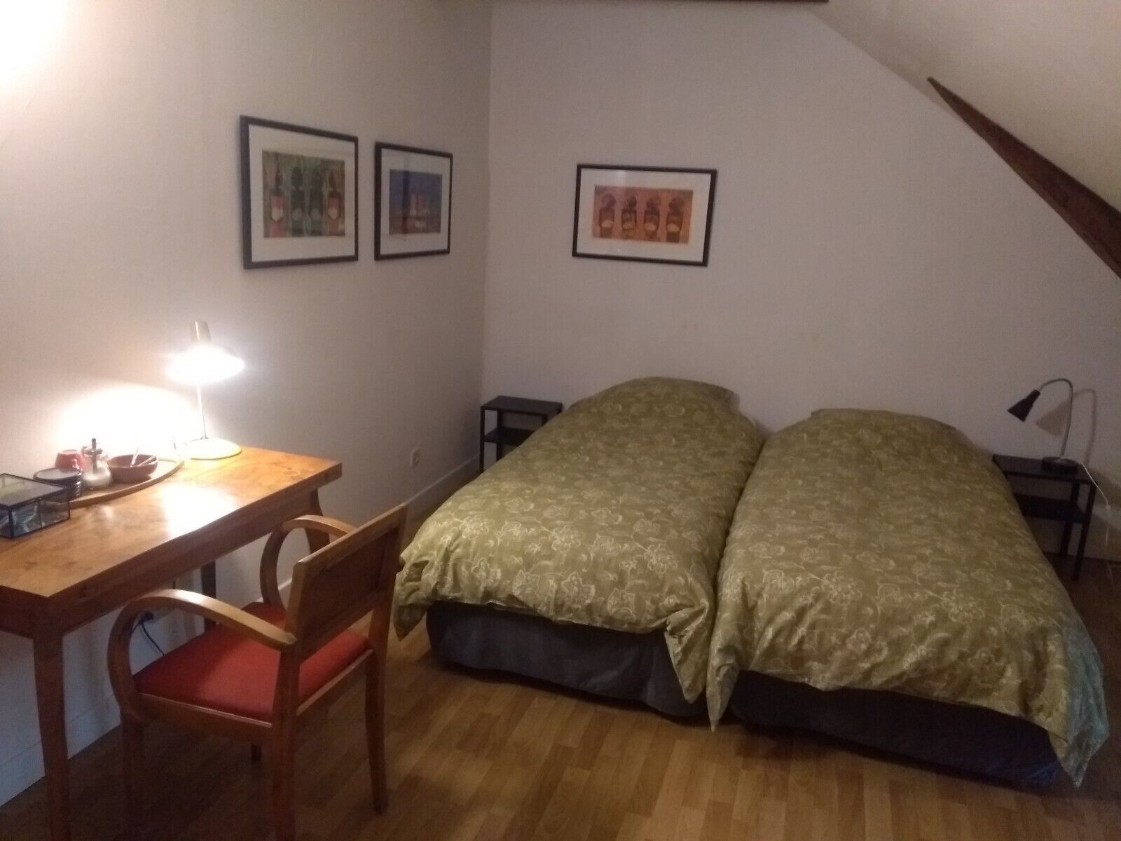 A small, cozy room with a wooden desk, a chair, a bed with a patterned bedspread, and two framed pictures on the wall.