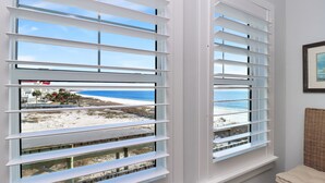 A room with two windows overlooking a beach, a wicker chair, and a framed picture on the wall.