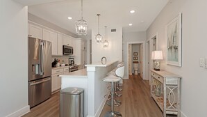 A modern kitchen with stainless steel appliances, a central island with bar stools, pendant lighting, and a hallway leading to a dining area.
