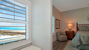 A bedroom with a view of the beach, a bedside table with a lamp, a chair, and a bed with patterned bedding.