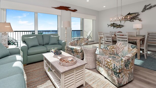 A spacious living room with a large window, a green sofa, a patterned armchair, a wooden coffee table, and a dining area with a table and chairs.