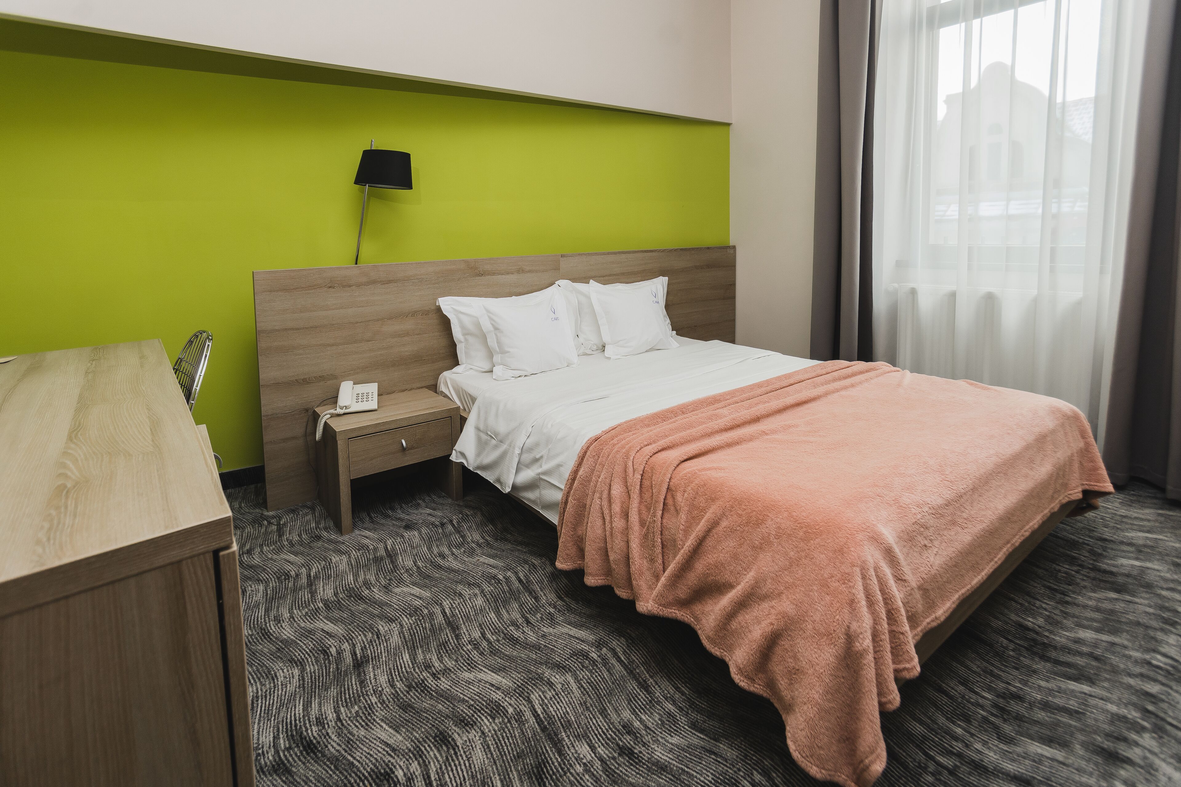 Una habitación de hotel con cama, escritorio, mesita de noche, lámpara y ventana con cortinas.
