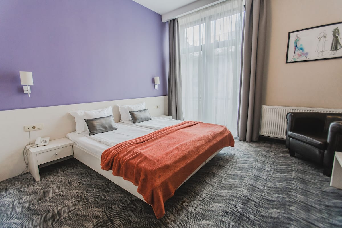 A hotel room with a bed, a nightstand with a phone, a chair, and a window with curtains.