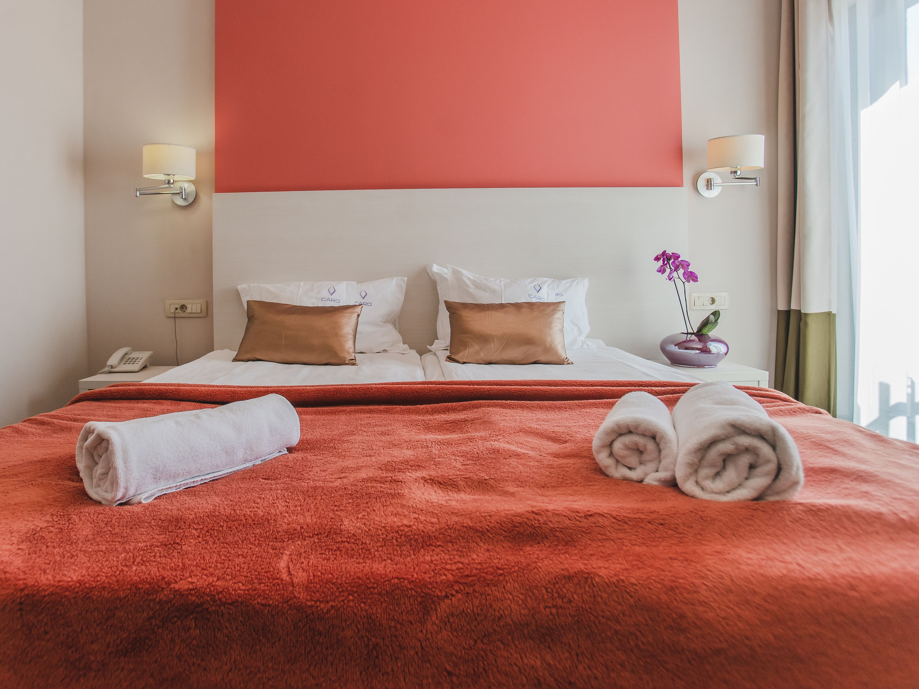 A hotel room with a large bed, two pillows, a red blanket, a white towel, a nightstand with a phone, and a wall-mounted lamp.