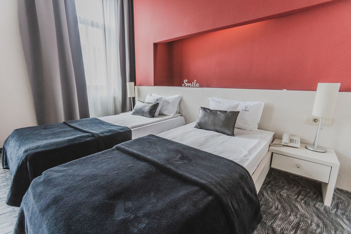 A hotel room with two beds, a red accent wall, a bedside table with a lamp, and a phone.