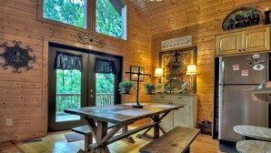 A rustic wooden interior with a large table and benches, a kitchen with a refrigerator, and a view of the outdoors through large windows. - Escape to Tranquility: Sleepy Hollow Cabin Retreat (Ellijay)