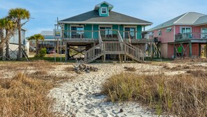 A beachfront house with a wooden deck, a staircase leading to the main entrance, and a sandy path leading to the door.