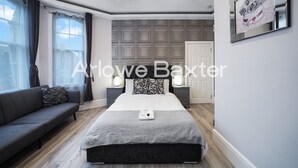 A modern bedroom with a bed, bedside tables, a sofa, and a large window with curtains. - Hackney Suites - Triple En-suite Room (F2) (Greater London)